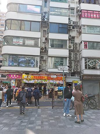 2）広場を右方向へ歩いていくと、横断歩道を渡った先に赤い看板のスナック屋さん（香港風スナックで、焼売などを売っている店）があるのが見えます。