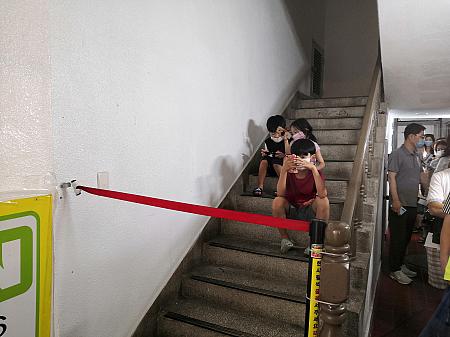 行列は通行人を妨げないように建物の通路に伸びていきます。待ちつかれた子供は列の横の階段でスマホゲームで時間つぶし。