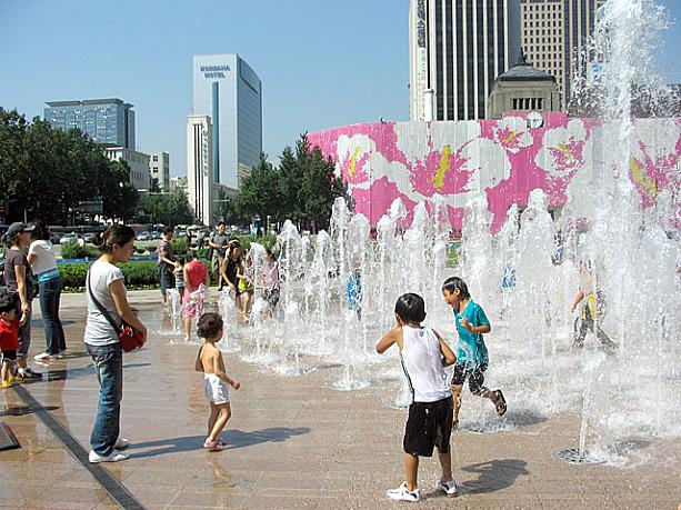 こちらは市庁にあるソウル広場。夏恒例となった噴水で遊ぶ子どもたちが楽しそうに遊んでいま〜す！後ろには８月１４日の「ほっとソウル」でお伝えした、市庁舎に施されたムクゲ柄の飾りが見られますね。
