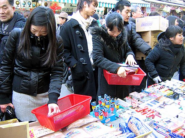 「どれにしようかな〜」と真剣にチョコを選ぶ女の子たちの後ろには、品切れしないようにと次々とチョコの入ったダンボールを運ぶお店の人や、「何だい？この騒ぎは？？」と見ている通りすがりのおじさん。