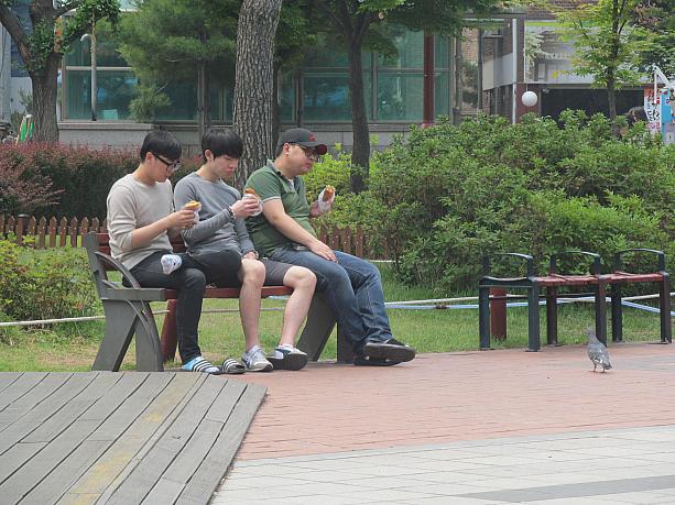 噴水の前のベンチで仲良く並んでお昼を食べている若い男性３人。鳩も食べたそう！？